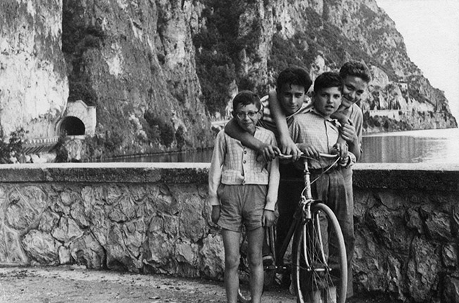 Old black and white photo of Nino as a young boy with his cousins and a bicycle with mountains and a lake in the background