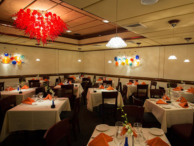 Photo of the private dining room with empty tables set with white table cloths and orange linen napkins. A striking Venetian glass chandelier is visible